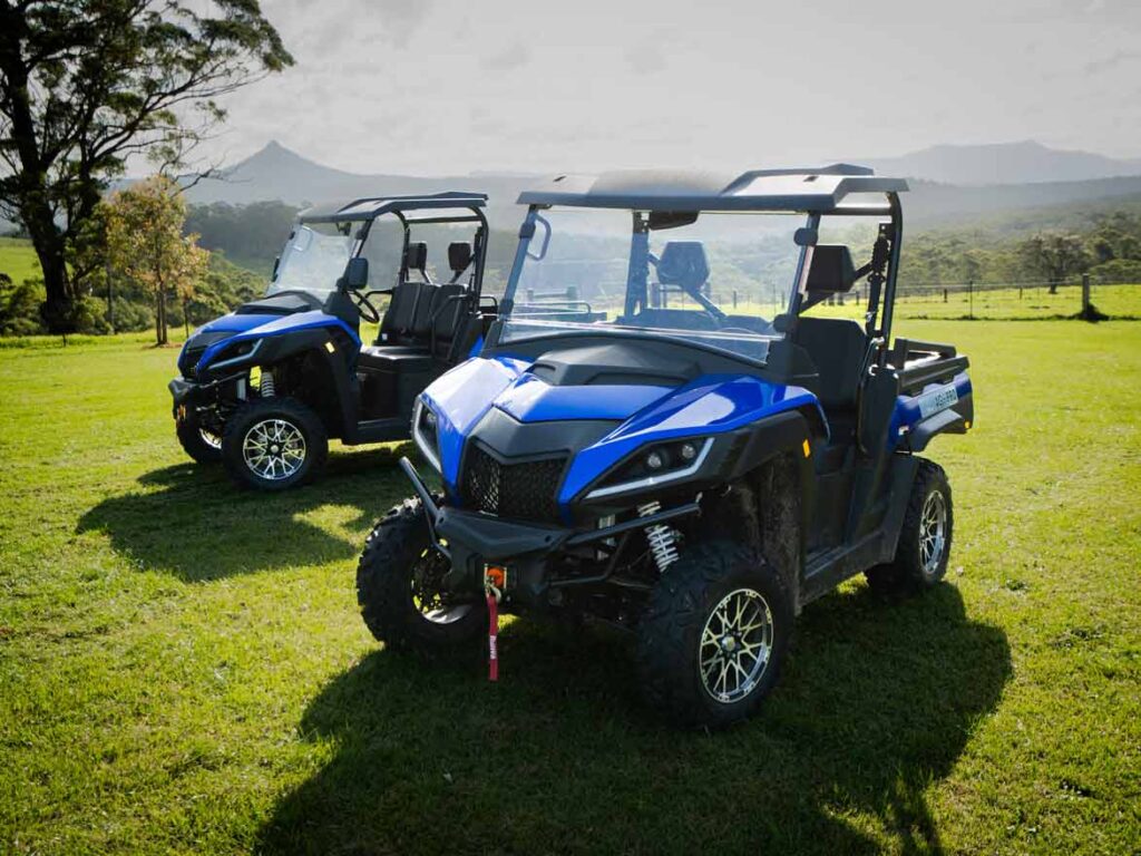 Zwei moderne, blaue UTVs mit Dach und Ladefläche, geparkt auf einer grünen Wiese vor einer hügeligen Landschaft.